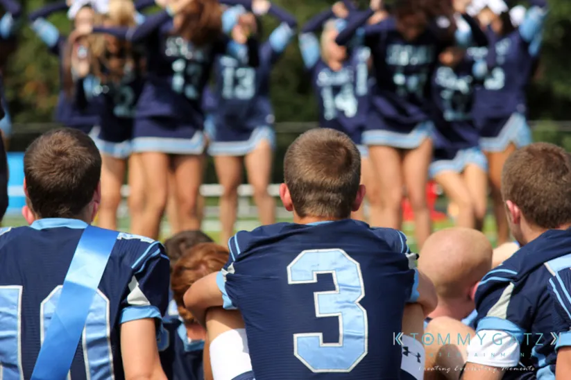 Football players in uniforms sit on sidelines watching cheerleaders perform on a field.