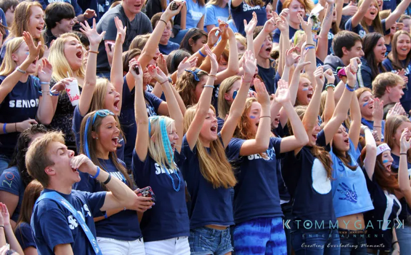 A group of excited students in blue senior shirts cheer enthusiastically in a crowd.