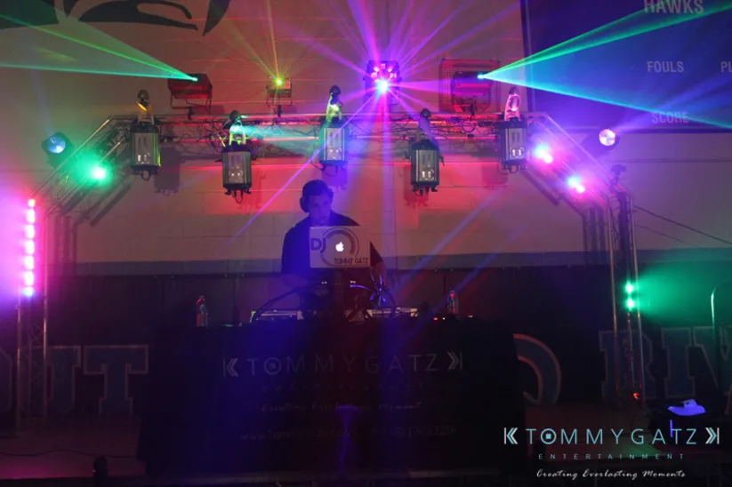 A DJ stands behind a booth with colorful lights and laser effects in a dark venue.