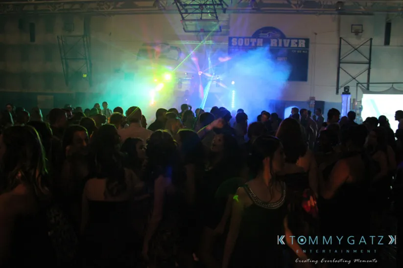 A group of people is dancing in a dimly lit indoor venue with colorful lights and fog effects.
