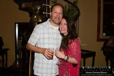 A man and woman smiling at a party, holding drinks, standing in front of a spiral staircase and decorative elements.