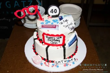 A two-tier birthday cake with a "40" topper, red glasses, and themed decorations. The cake reads "Happy 40th Birthday Orin" and includes weekday initials.
