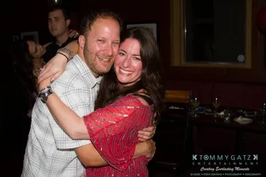 A smiling couple embraces in a dimly lit room, posing for the camera. Other people are visible in the background.