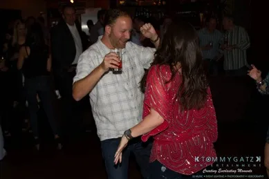 A man and a woman dance together at a social event. The man holds a drink, and both appear to be enjoying the moment.