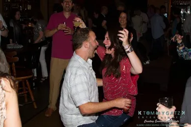 A man and woman laugh and dance in a lively, crowded bar with people enjoying drinks in the background.
