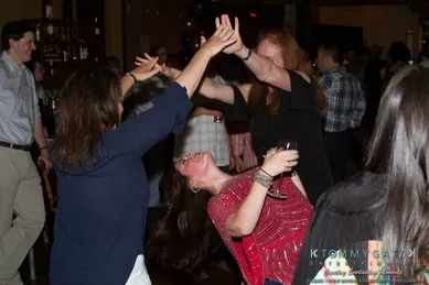 People dancing energetically at a party, with one woman in a red dress leaning back while holding a drink. Others around her are engaging in lively dance moves.