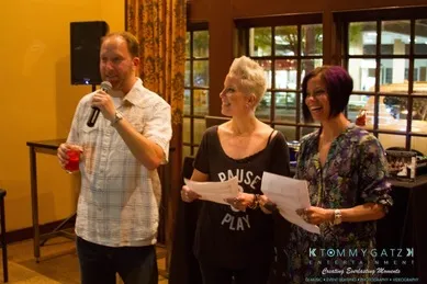 Three people stand indoors, two holding papers and one speaking into a microphone. They appear to be presenting or hosting an event.