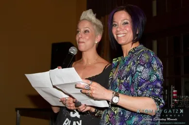 Two people stand side by side, each holding papers. One is speaking into a microphone, while the other smiles.