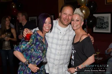 Three people smiling and posing together at a party. One person is holding a microphone. Balloons and other guests are visible in the background.