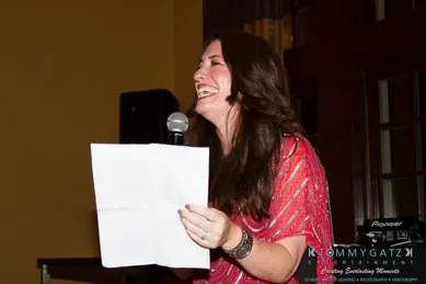 A woman in a red top is speaking into a microphone, smiling, and holding a sheet of paper.