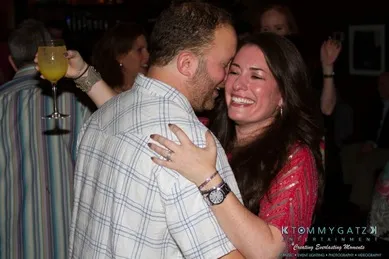A couple embraces and laughs at a social gathering. The man holds a drink in his hand.