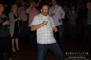 A man in a plaid shirt dances with a drink in hand at a party, surrounded by smiling people.