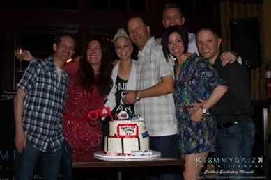 A group of people standing around a decorated cake, smiling for the camera in a dimly lit room.