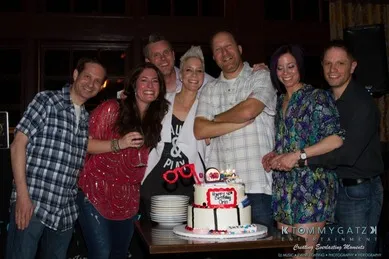 A group of people standing around a table with a cake. Some are posing with their arms around each other, and the room is dimly lit.