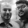 Two men in shirts and ties, standing close together, looking at the camera. Image in black and white.