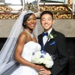 Bride in a white dress and groom in a dark suit with blue accents pose together, smiling, holding a bouquet, in front of an ornate backdrop.