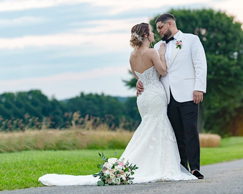 Bride in a white gown and groom in a white jacket pose together outdoors, with a bouquet on the ground.