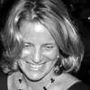 Black and white photo of a person with shoulder-length hair smiling and looking downward, wearing a dark top and necklace.