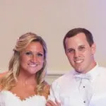 A couple in formal attire smiles at the camera; the woman is in a white dress and the man in a white shirt with a bow tie.