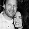 A black and white photo of a smiling couple, with a man on the left wearing a checkered shirt and a woman on the right with long dark hair.