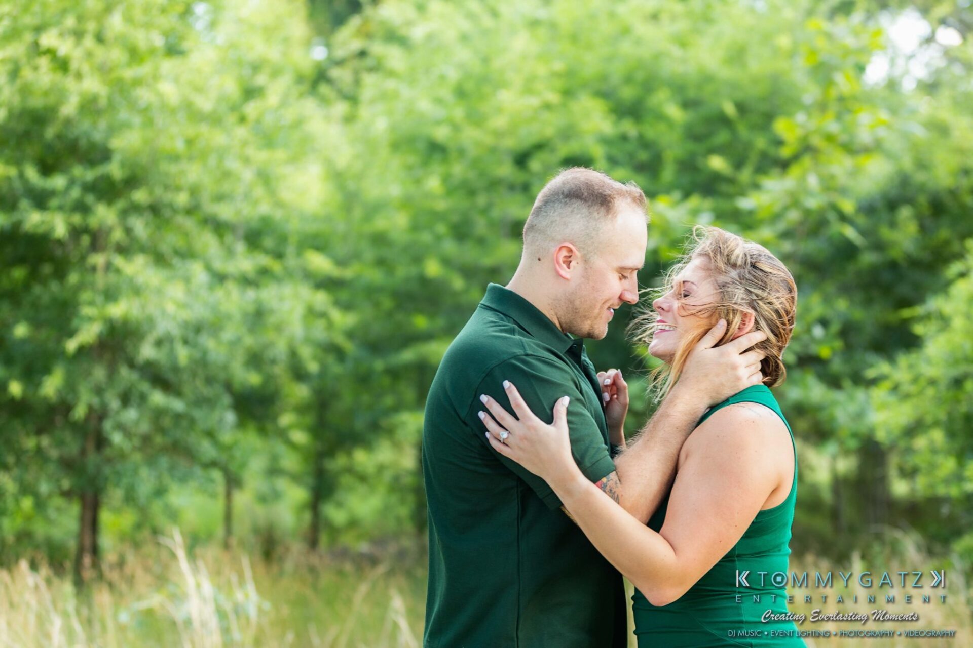 A couple stands close together, smiling at each other, in a lush green setting.