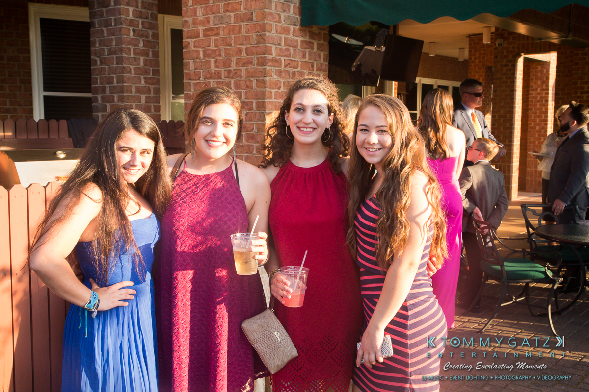 Garbis-Grad-Edits-lowres-Watermark-106 - TOMMY GATZ ENTERTAINMENT Four women in colorful dresses smile and hold drinks at an outdoor gathering with brick walls and a patio.