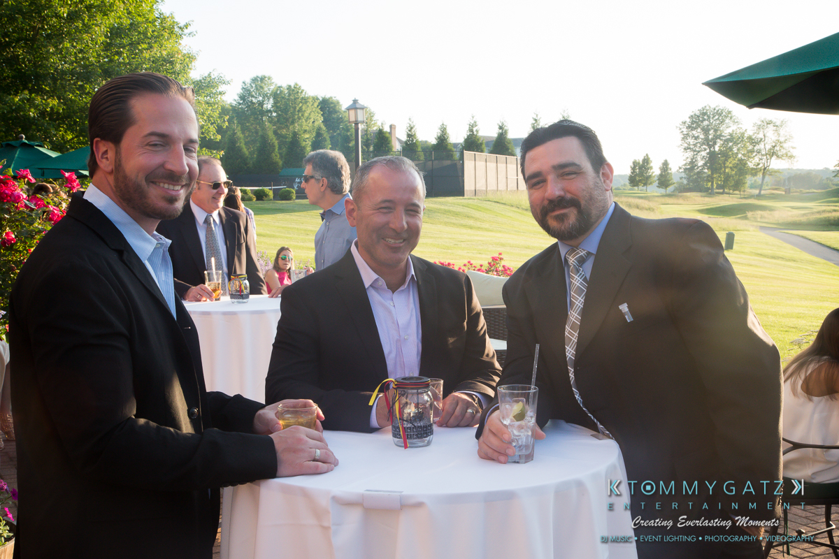 Garbis-Grad-Edits-lowres-Watermark-107 - TOMMY GATZ ENTERTAINMENT Three men in suits stand around a cocktail table outdoors, holding drinks and smiling. The setting is sunny with greenery and a building in the background.