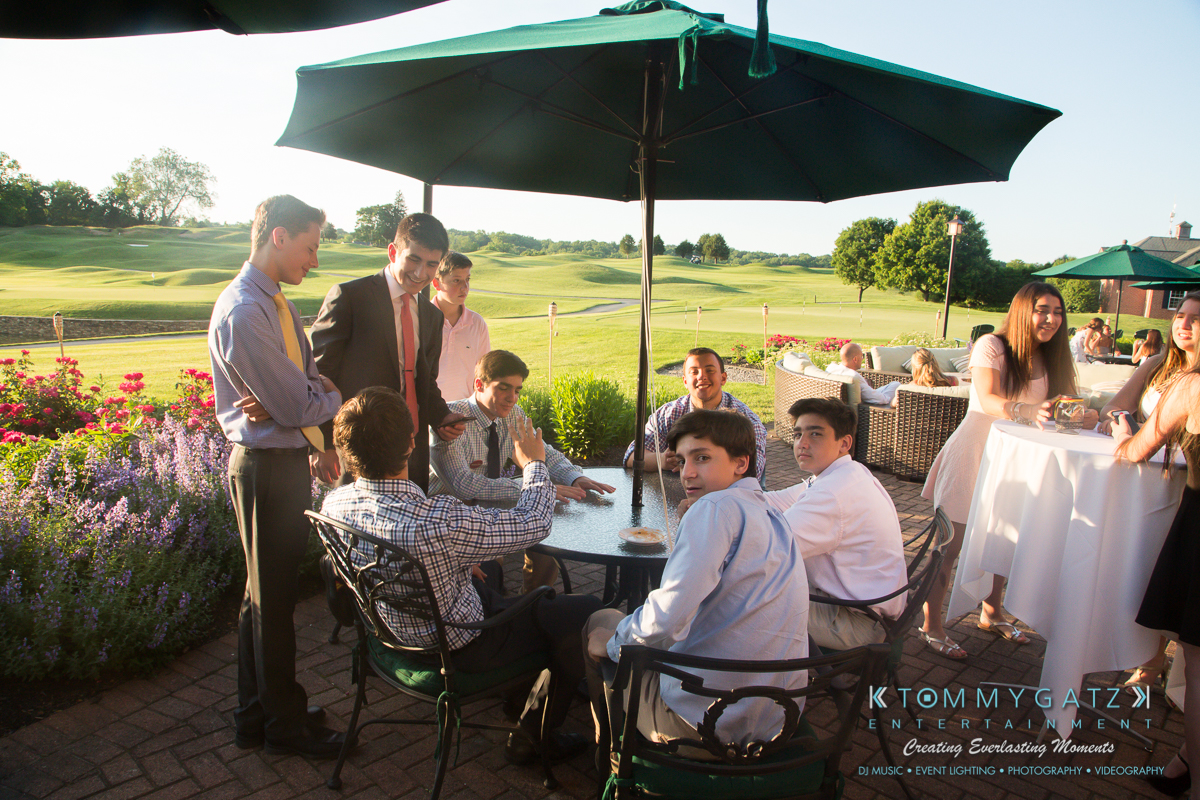 Garbis-Grad-Edits-lowres-Watermark-110 - TOMMY GATZ ENTERTAINMENT A group of young men seated around a patio table under an umbrella at an outdoor event, with others standing and mingling nearby. The setting is a sunny golf course.