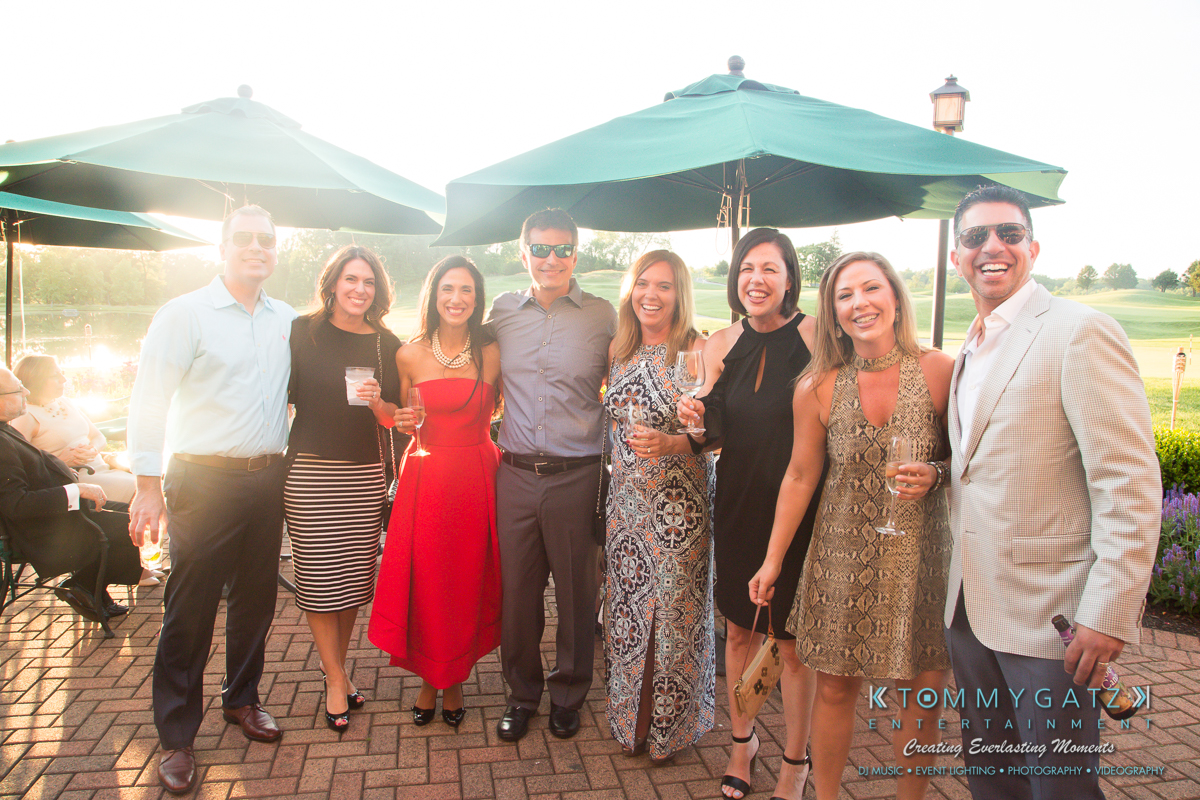 Garbis-Grad-Edits-lowres-Watermark-124 - TOMMY GATZ ENTERTAINMENT A group of people standing together outdoors, smiling, with beverages in hand. They are dressed in semi-formal attire. Large green umbrellas are in the background.
