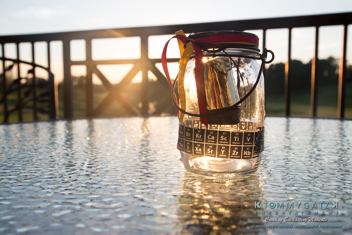 Garbis-Grad-Edits-lowres-Watermark-133 - TOMMY GATZ ENTERTAINMENT A glass jar with a periodic table design and ribbons sits on a glass table. Sunlight shines through, with a wooden fence visible in the background.
