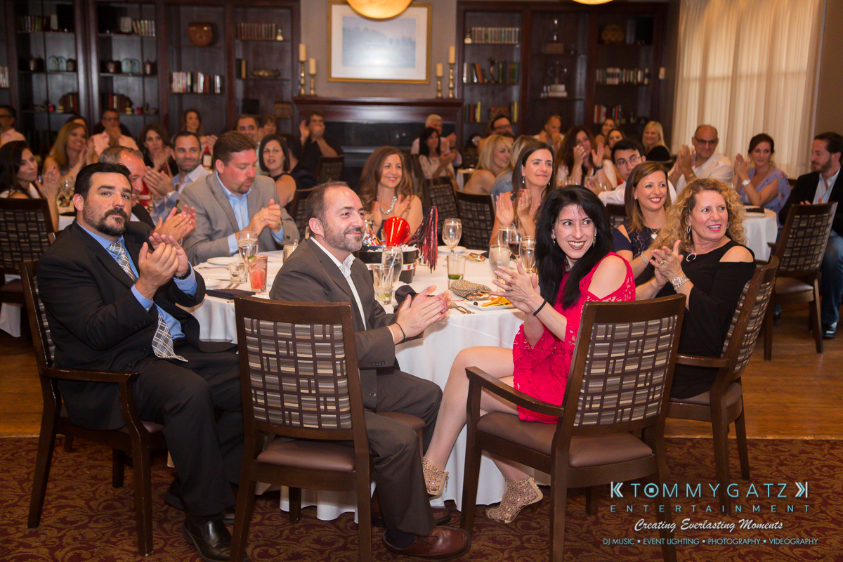 Garbis-Grad-Edits-lowres-Watermark-165 - TOMMY GATZ ENTERTAINMENT A group of people seated at round tables in a dining room clapping, with a large bookshelf and framed picture in the background.