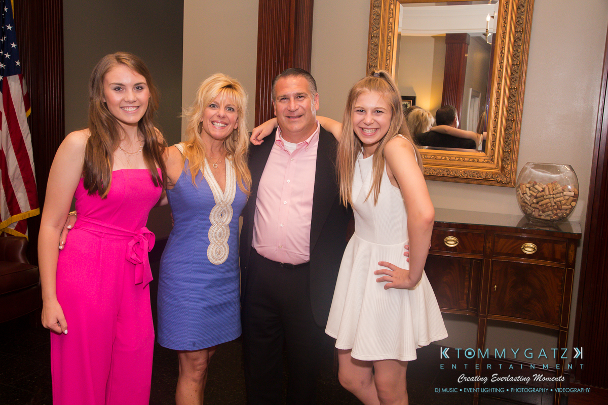 Garbis-Grad-Edits-lowres-Watermark-232 - TOMMY GATZ ENTERTAINMENT A group of four people stand together in a room with a U.S. flag and decorative mirrors in the background, smiling at the camera.
