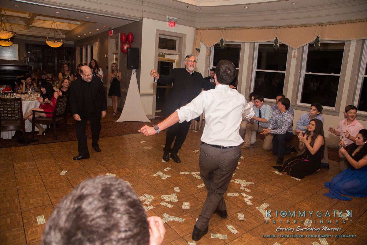 Garbis-Grad-Edits-lowres-Watermark-272 - TOMMY GATZ ENTERTAINMENT Two men dance near a group seated on the floor, with dollar bills scattered around. A seated crowd watches in a reception-style venue.