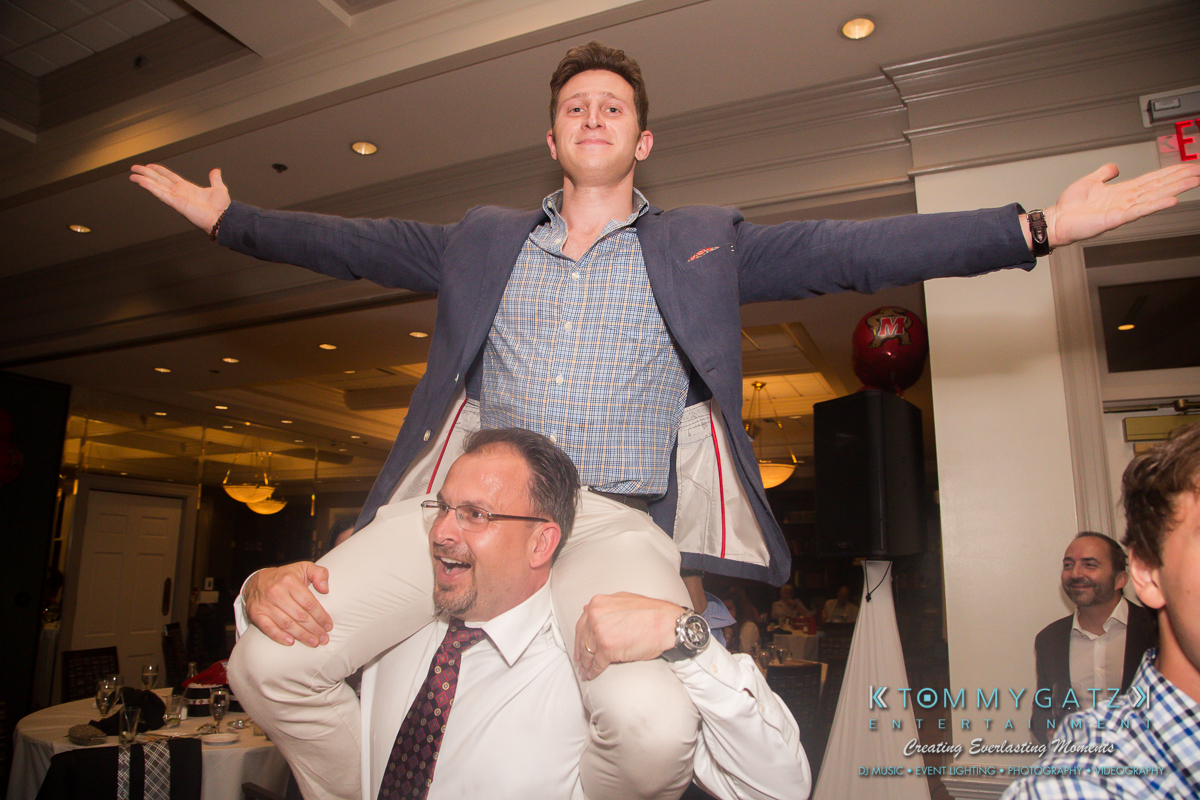 Garbis-Grad-Edits-lowres-Watermark-302 - TOMMY GATZ ENTERTAINMENT A man in a suit carries another man on his shoulders at an indoor event. The man on top has his arms spread wide, and people are seated around them.