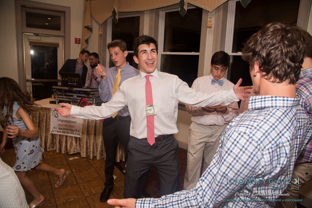 Garbis-Grad-Edits-lowres-Watermark-326 - TOMMY GATZ ENTERTAINMENT A group of young people dancing and socializing in a room with large windows. One person in a white shirt and pink tie appears to be animatedly spreading his arms.