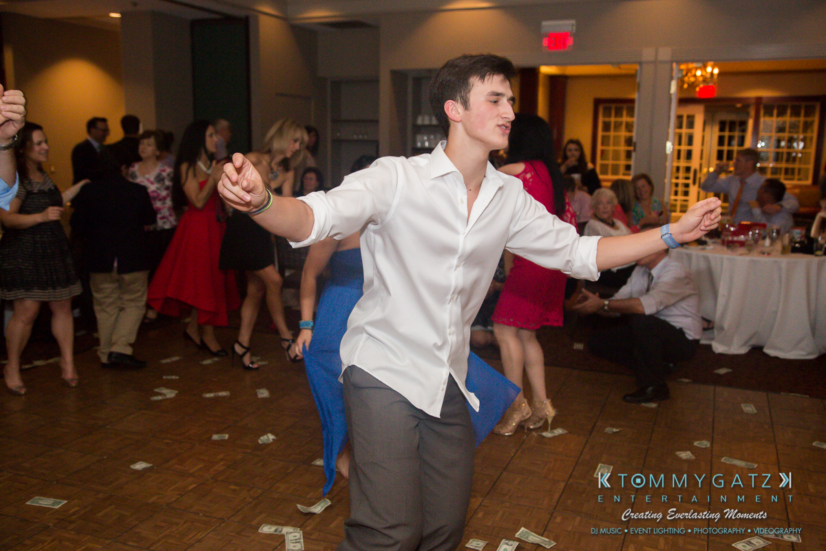 Garbis-Grad-Edits-lowres-Watermark-431 - TOMMY GATZ ENTERTAINMENT Young man dancing energetically on a dance floor surrounded by scattered dollar bills, with people seated and standing in the background.