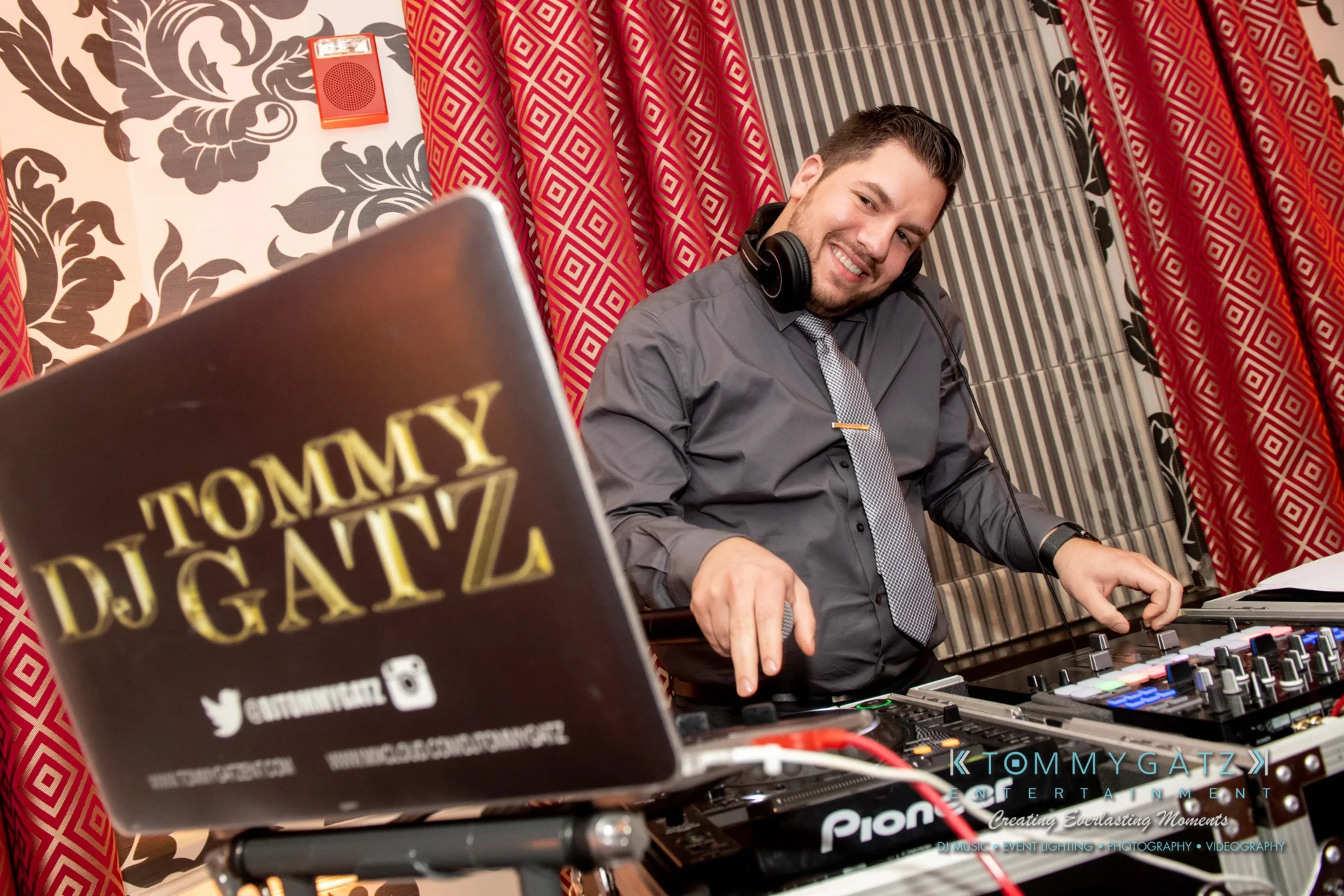 A DJ in a gray shirt and black tie stands behind a DJ mixer. He is smiling with headphones around his neck. The laptop on the table displays "Tommy DJ Gatz.
