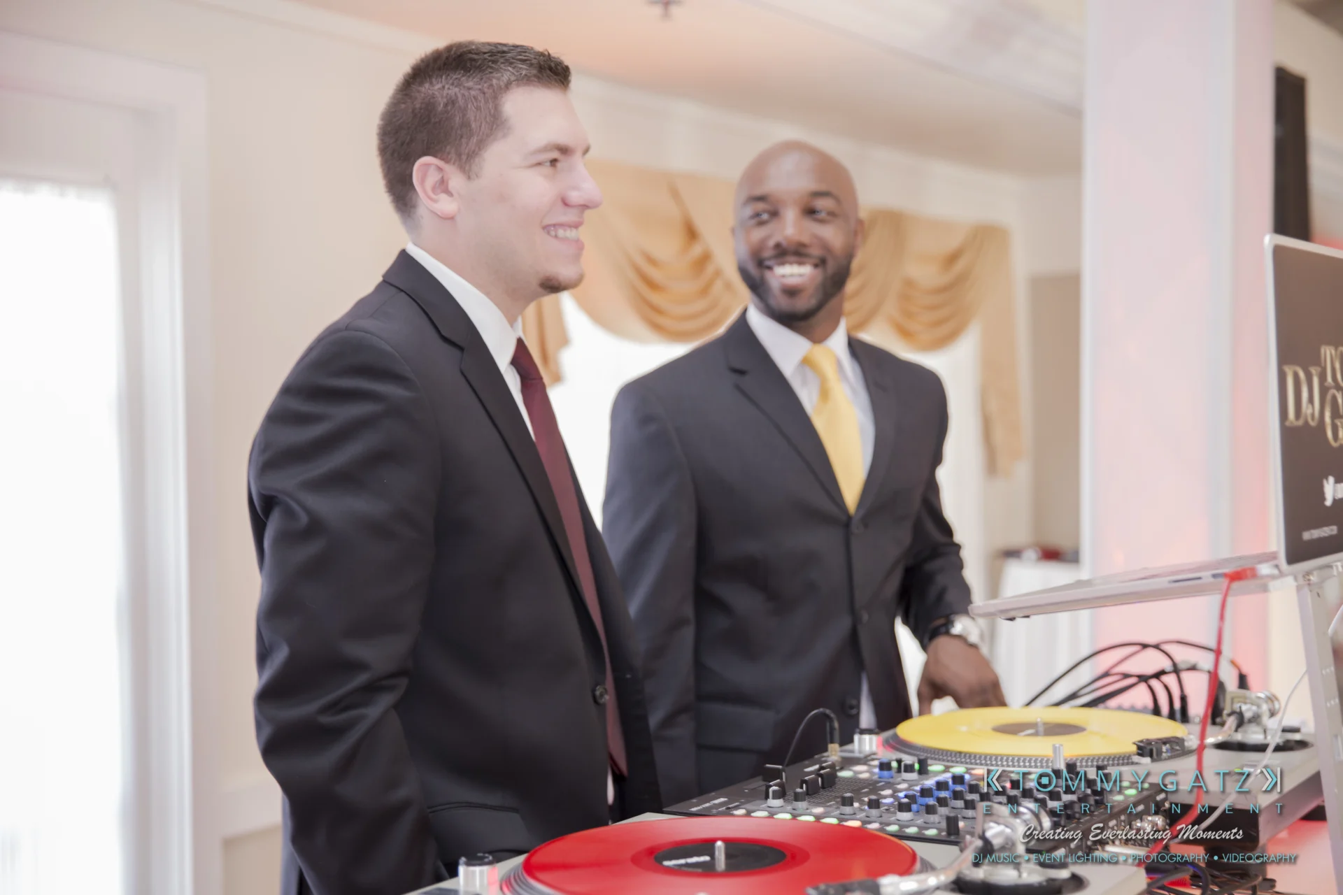 Two men in suits smiling near a DJ booth with red and yellow turntables in an event setting.