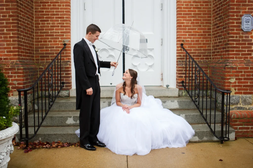 wedding-photographer-annapolis-tommy-gatz-entertainment - TOMMY GATZ ENTERTAINMENT A bride sits on steps in a white gown holding an umbrella as a groom stands beside her in a tuxedo, outside a brick building with white doors.