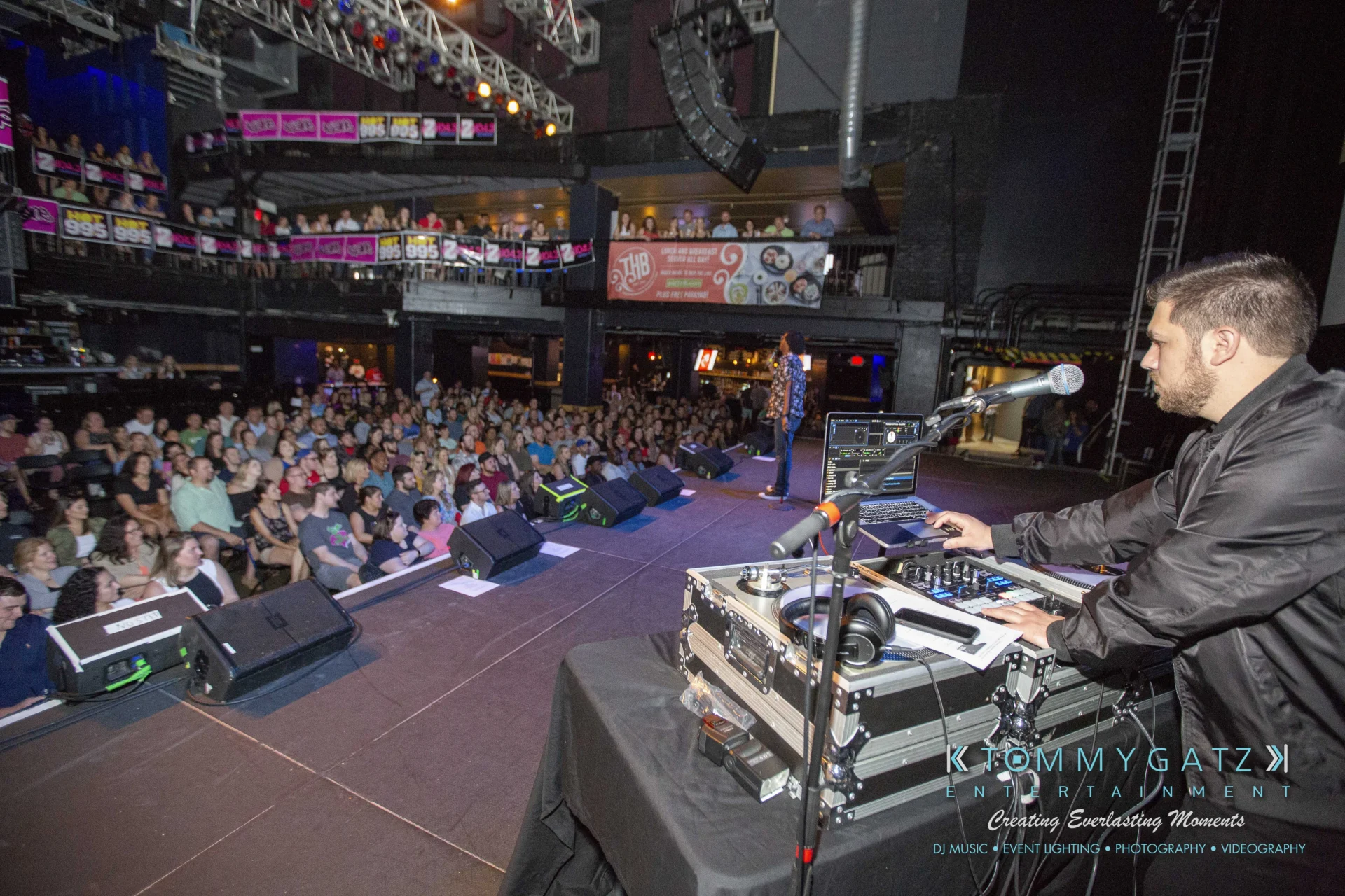 DJ performing on stage, audience in background at a lively indoor event venue.