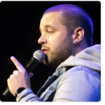 Person with a beard holding a microphone, speaking, and gesturing with their hand. Wearing a denim jacket and hoodie. Black background.
