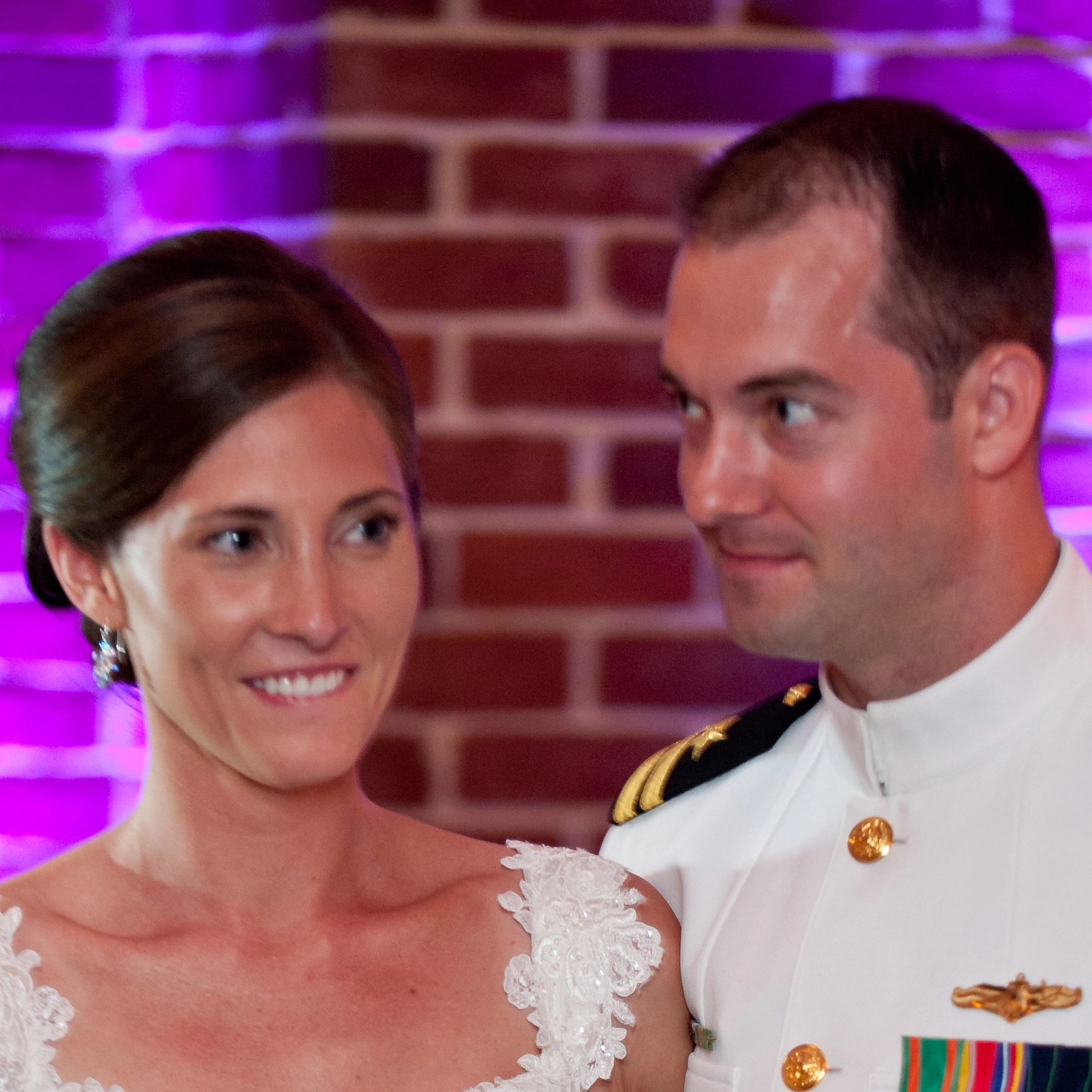 A woman in a wedding dress and a man in a military uniform stand together, smiling. Brick wall backdrop.