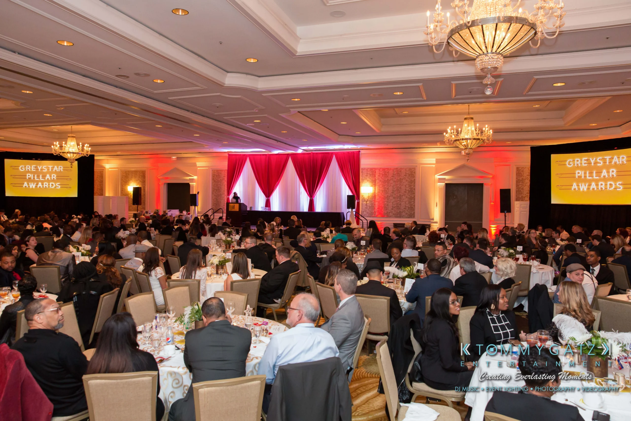 A large banquet hall filled with people seated at tables. Two screens display "Greystar Pillar Awards." A stage with red curtains is visible at the front.