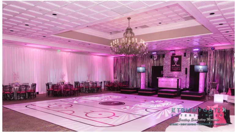 An elegant event hall with a pink-lit ceiling, large chandelier, and decorated stage. The floor features a custom hockey rink design with combined logos "DYLAN" and "TOMMY CATZ ENTERTAINMENT.