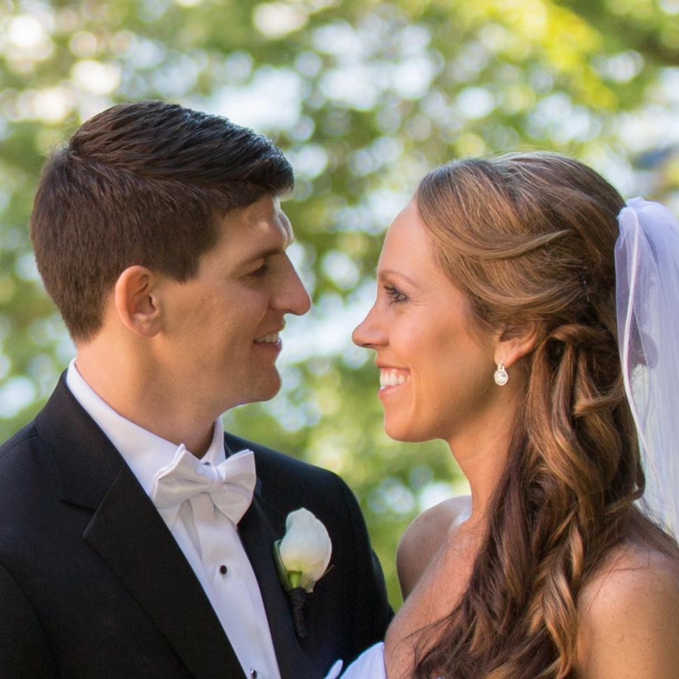 A couple stands outdoors on their wedding day, facing each other and smiling. The groom wears a black tuxedo, and the bride wears a white wedding dress with a veil.