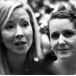 Two women in a black and white photo, one with long hair speaking, the other with shorter hair looking at the camera, blurred background.