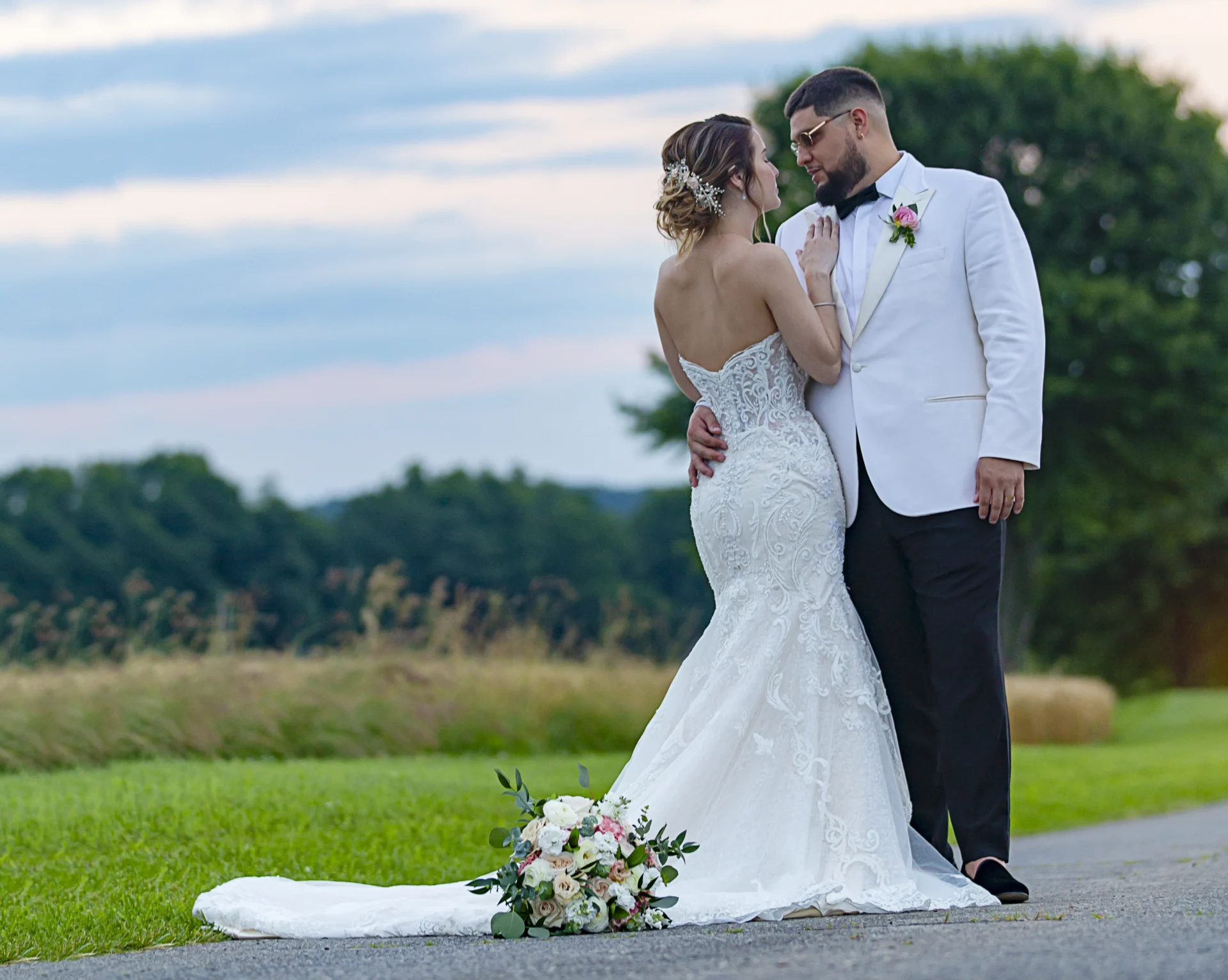 Couple in wedding attire embrace on a path with a bouquet on the ground. Backdrop of green grass and trees under a blue sky.