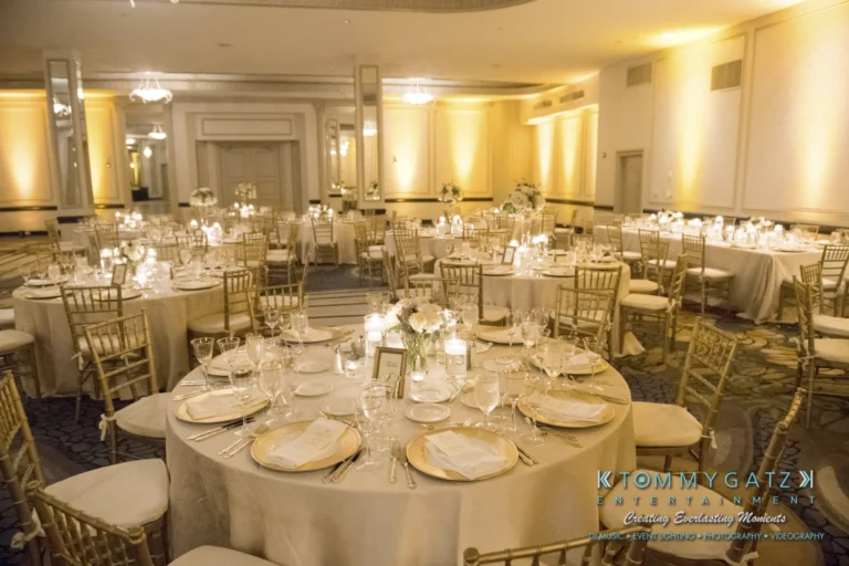 Elegant banquet hall with round tables set for an event, featuring white tablecloths, gold chairs, and dim lighting. Floral centerpieces and candles adorn the tables. Logo in the corner.