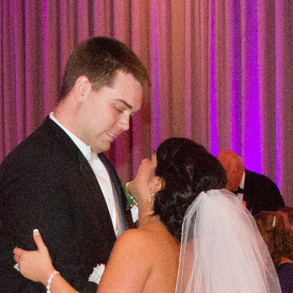 A couple dances together in formal attire, with the woman wearing a wedding veil. A curtain with pink and purple lighting is in the background.