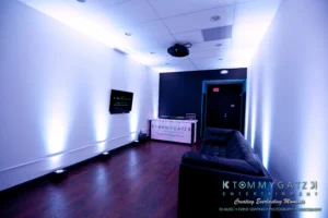 Dimly lit office lobby with a dark couch, television on the wall, and a reception desk labeled "Tommy Gatz Entertainment." Purple and blue lights illuminate the space.
